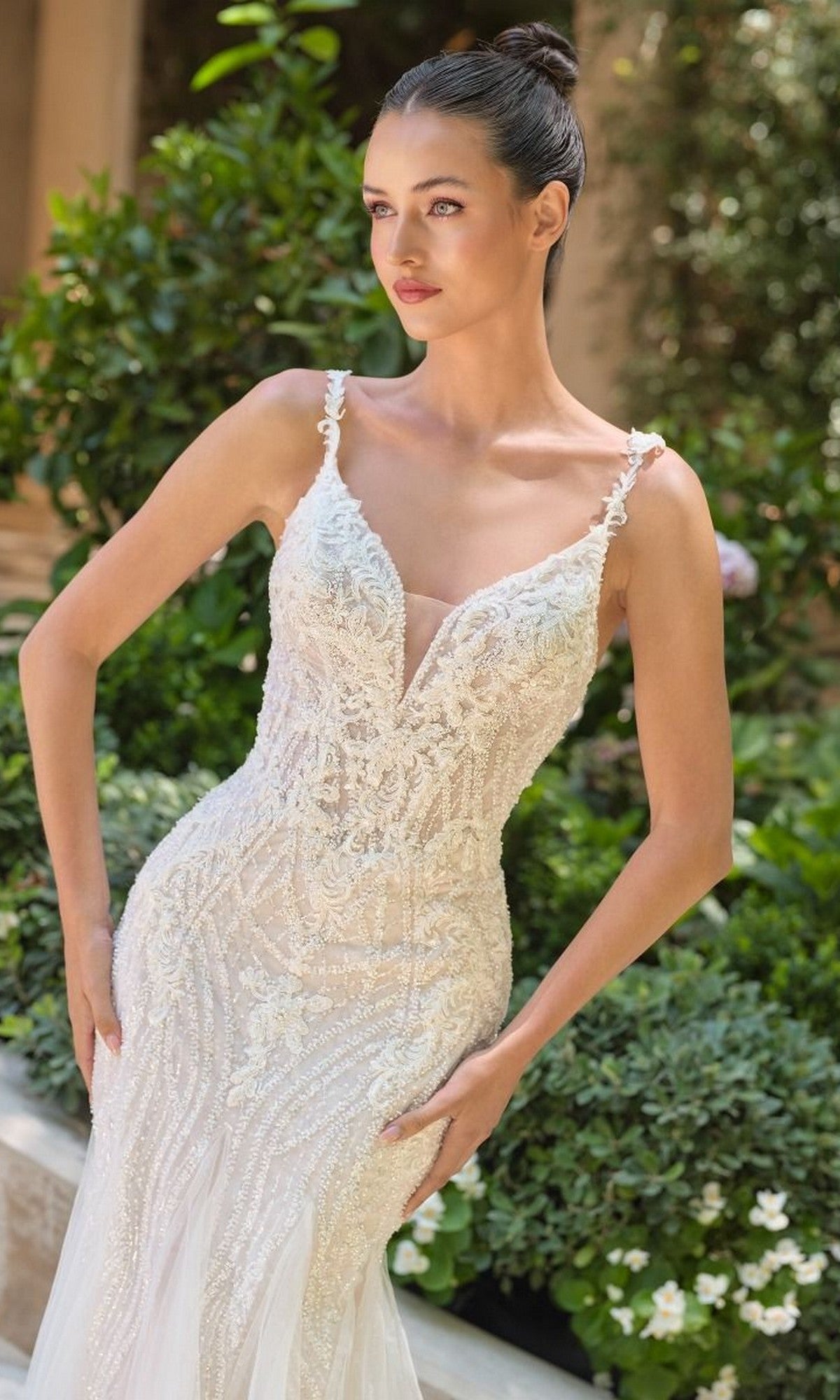 A woman stands outdoors in the Long Wedding Dress: Andrea and Leo WL066—a fit-and-flare gown with intricate beaded embroidery, delicate white lace, thin straps, and a fitted silhouette—surrounded by lush greenery.